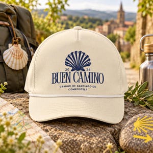 Peut inclure: Une casquette de baseball couleur crème avec un motif bleu foncé représentant une coquille Saint-Jacques au-dessus du texte 2026 BUEN CAMINO CAMINO DE SANTIAGO DE COMPOSTELA. La casquette présente une calotte structurée et un cordon blanc.