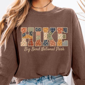 Puede incluir: Una sudadera marrón de cuello redondo con la palabra HIKE en letras grandes de bloque rellenas con un mosaico de patrones florales y de cuadros en tonos azul, naranja, verde y blanco. Debajo, el texto Big Bend National Park está impreso.