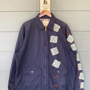 May include: Navy blue jacket with a zip-up front, two front pockets, and decorative patches. The patches are square with a plaid pattern and a light blue design. The jacket is hanging on a wooden hanger against a light blue background.