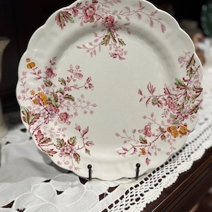 May include: A round ceramic plate with a scalloped edge featuring a cream-colored base and a delicate floral pattern in shades of pink, green, and yellow. The botanical design is arranged around the perimeter, leaving the center of the plate clear.