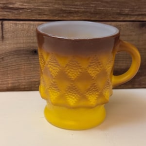 May include: A vintage yellow and brown glass mug with a textured diamond pattern. The mug has a rounded handle and a white interior. The base and handle are yellow, with a gradient from brown to yellow on the body.