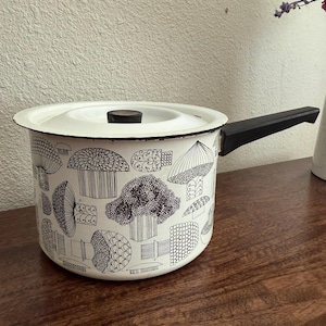 May include: White enamel saucepan with a black handle and lid. The pot is decorated with a black and white mushroom pattern. The pot is sitting on a wooden surface.