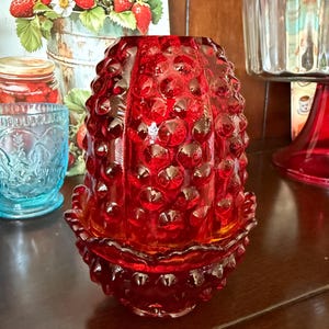 May include: A red glass candle holder with a hobnail pattern. The candle holder has a flared base and a scalloped edge. The glass is a deep red colour. The candle holder is likely vintage or antique.