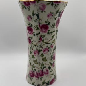 May include: A white ceramic vase with a flared top, adorned with a pattern of pink roses and green leaves. The rim is trimmed with gold. The vase is approximately 8 inches tall.