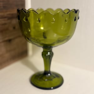 May include: A green glass pedestal bowl with a scalloped rim and teardrop-shaped details. The bowl has a rounded base and a stem. The glass has a slight green tint, and the bowl is likely vintage.