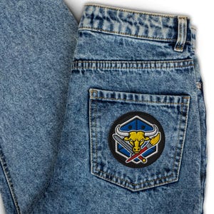 May include: Light blue denim jeans with a black patch on the back pocket. The patch features a yellow bull with white horns, crossed swords, and a red paintbrush, set against a blue and white geometric design.