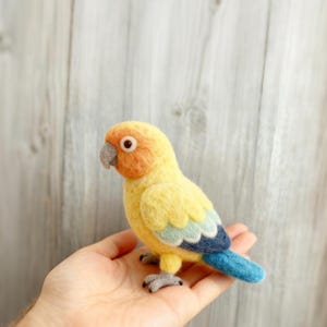 May include: A handcrafted felt bird figurine in yellow, orange, and blue hues. The bird has a gray beak and feet, and is perched on a hand. The bird's body is primarily yellow, with orange on its head and blue accents on its wings and tail.