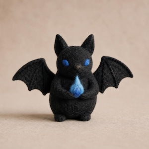 Può includere: Una piccola statuetta di pipistrello nera in lana infeltrita, con orecchie a punta e ali spiegate. La creatura ha occhi blu brillante e tiene tra le zampe anteriori una piccola forma di fiamma blu con un effetto luminoso.