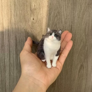 Op de afbeelding: Een klein naaldgevilte kattenfiguurtje met donkergrijze en witte vacht. De kat heeft een witte borst, witte pootjes en een wit gezicht met een roze neus, donkere ogen en lange witte snorharen. Het figuurtje heeft een getextureerd, pluizig uiterlijk.