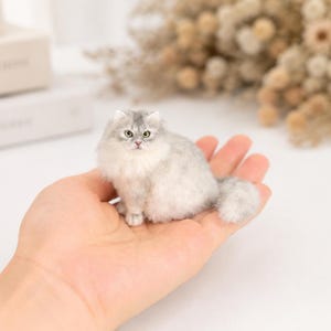 May include: A miniature figurine of a fluffy long-haired cat with silver and white fur, pointed ears, and green eyes. The small cat is depicted in a sitting position with its tail curled slightly to the side, showcasing detailed fur texture.