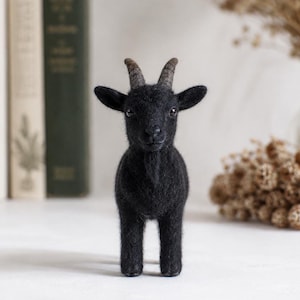 May include: A needle-felted black goat figurine with a fuzzy texture, small pointed ears, two curved greyish-brown horns, and a small beard. The goat stands on four legs, featuring dark eyes and a detailed snout, crafted from wool fibers.