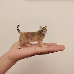 Può includere: Una dettagliata statuetta di gatto in miniatura, principalmente beige con segni marroni più scuri, è appoggiata su una mano. Il gatto ha un aspetto strutturato, con una coda a strisce e dettagliati tratti del viso. È un piccolo oggetto fatto a mano.