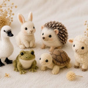 Puede incluir: Una colección de seis figuras de animales de lana fieltrada que incluye un pato blanco, un conejo blanco, un erizo marrón y blanco, una rana verde, una pequeña tortuga marrón y crema, y un cordero color crema, dispuestos con piñas y flores.