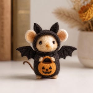 May include: A needle-felted mouse figurine wearing a black bat costume with pointed ears and wings. The mouse has light beige fur, black eyes, and a thin tail, and it is holding a small orange felt pumpkin bucket with a black jack-o'-lantern face.