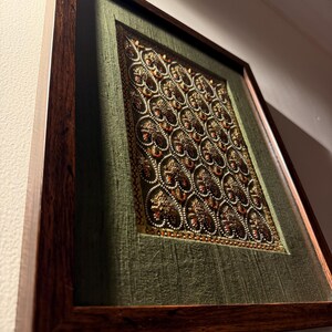 May include: A rectangular wall art piece featuring a dark brown wooden frame surrounding a textured olive green fabric background. The center displays an intricate embroidery pattern of repeating gold and copper heart shapes with bead accents.