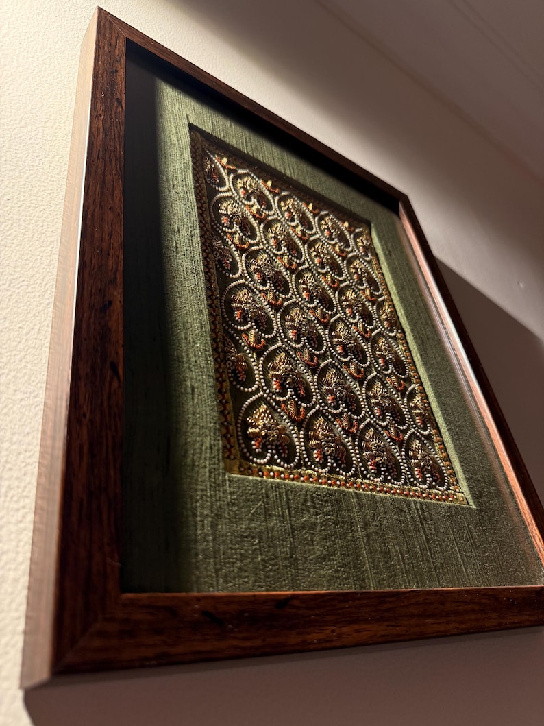 May include: A rectangular wall art piece featuring a dark brown wooden frame surrounding a textured olive green fabric background. The center displays an intricate embroidery pattern of repeating gold and copper heart shapes with bead accents.