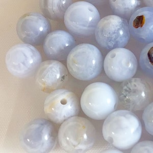 May include: A collection of smooth, round, light blue agate beads with white and brown markings. The beads are arranged in a random pattern on a white surface.