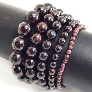 May include: A stack of seven garnet gemstone bracelets with varying bead sizes. The bracelets are made of dark red, round beads.