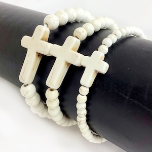May include: Three white beaded bracelets with white cross charms. The bracelets are layered on a black surface.