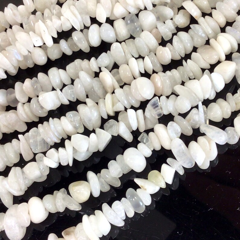 May include: A strand of white moonstone beads, featuring a mix of smooth, rounded, and flat, irregular shapes. The beads are strung together on a clear thread.