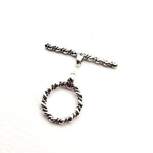 May include: A silver-toned toggle clasp set, featuring a twisted rope design. The set includes a circular ring and a bar, both with a textured, braided appearance. The clasp is designed for jewelry making.