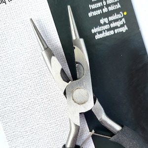 May include: Close-up of round nose pliers with a silver metal finish and black cushioned grips. The pliers are designed for precision work, featuring pointed tips and a sturdy construction. The background includes text in a foreign language.