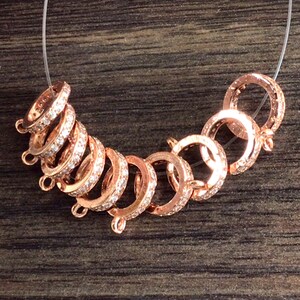 K&ouml;nnte beinhalten: Ein Satz von acht ros&eacute;goldfarbenen Metallverschl&uuml;ssen mit klaren Kristall-Akzenten. Jeder Verschluss ist rund mit einer kleinen Schlaufe an jeder Seite.
