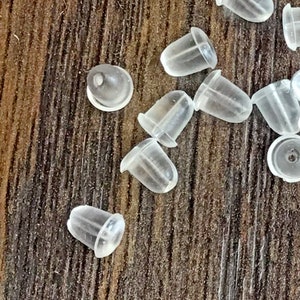 May include: A close-up of a group of clear plastic earring backs. The backs are small and round, with a slight indentation in the centre. They are scattered on a wooden surface.