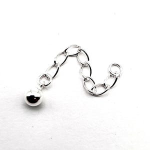 May include: A silver-colored chain extender with a small ball at one end. The chain is composed of oval links and is designed to add length to necklaces or bracelets. The extender is shown against a white background.