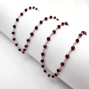 May include: A delicate necklace featuring small, faceted, deep red beads strung on a silver chain. The beads are evenly spaced, creating a continuous line. The necklace is displayed against a white background, highlighting the contrast of colors.