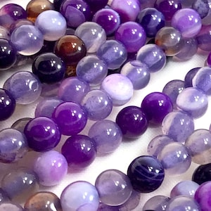 May include: A strand of purple agate beads with varying shades of purple and white. The beads are round and smooth, and they have a natural, polished finish.