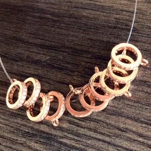 K&ouml;nnte beinhalten: Ein Satz von acht ros&eacute;goldfarbenen Metallringen mit klaren Kristall-Akzenten. Die Ringe sind f&uuml;r die Verwendung als Schmuckherstellungskomponenten konzipiert.
