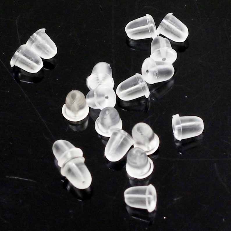 May include: A close-up of a pile of clear plastic earring backs. The backs are small and round, with a hole in the centre. They are designed to be used with stud earrings.