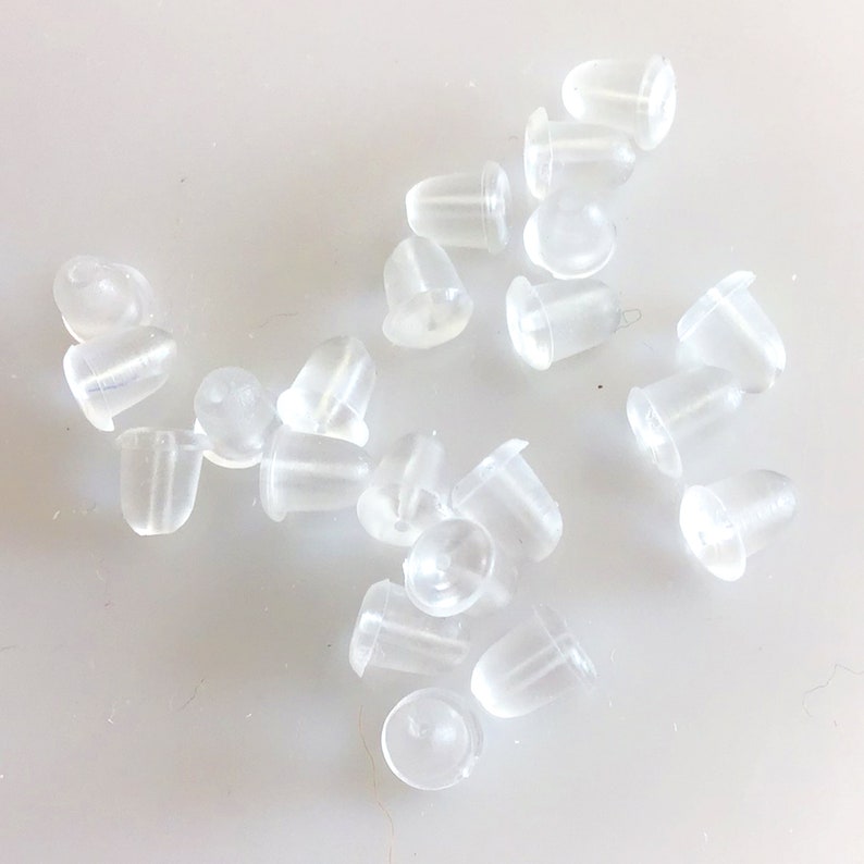 May include: A close-up of a pile of clear plastic earring backs. The backs are small and round, with a small hole in the centre.