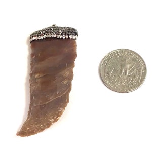 May include: A pendant with a brown, irregular shape, possibly stone or agate, topped with a silver-colored cap encrusted with small, sparkling black and clear stones. A U.S. quarter coin is next to the pendant for size comparison.