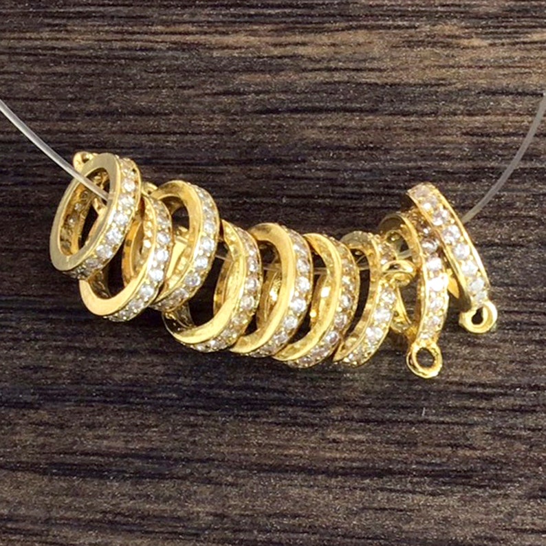 K&ouml;nnte beinhalten: Eine Gruppe von goldfarbenen Metallringen mit klaren Kristallverzierungen. Die Ringe sind &uuml;bereinander gestapelt und haben eine glatte, polierte Oberfl&auml;che.