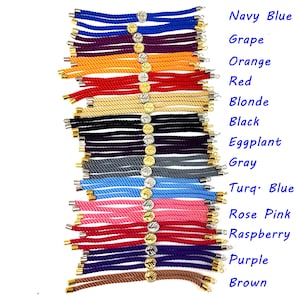 May include: A collection of 15 colorful rope bracelets with gold accents. The bracelets are arranged vertically, with the colors ranging from navy blue to brown. Each bracelet has a small gold charm attached to it.