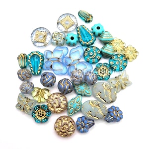 May include: A collection of assorted glass beads in shades of blue, turquoise, and gold. The beads feature various shapes including butterflies, flowers, stars, moons, and geometric patterns, with some displaying metallic gold detailing on the surfaces.