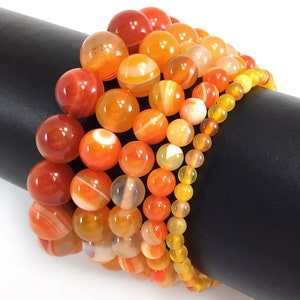 May include: A set of six bracelets made with orange and yellow agate beads. The bracelets are stacked on a black surface.