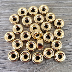 Pu&ograve; includere: Un gruppo di 25 perline di metallo dorate con un design ad anello rialzato. Le perline sono disposte in modo casuale su una superficie di legno.