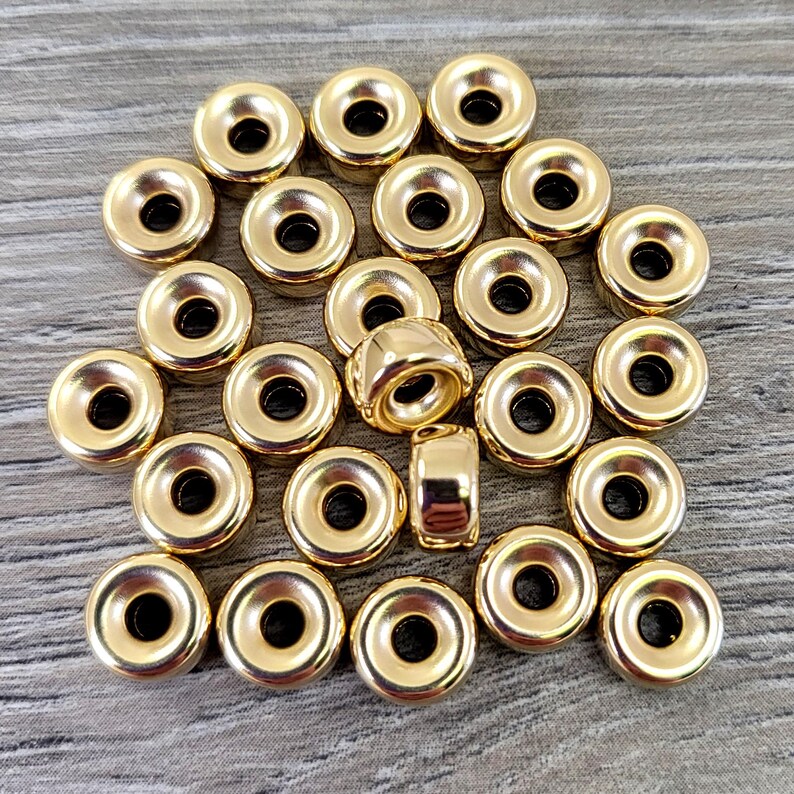 Pu&ograve; includere: Un gruppo di 25 perline di metallo dorate con un design ad anello rialzato. Le perline sono disposte in modo casuale su una superficie di legno.