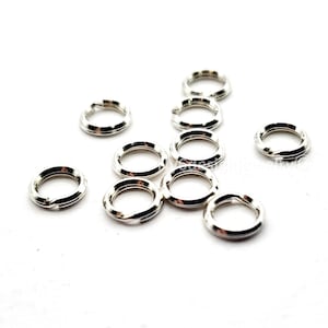 May include: A collection of small, silver-colored split rings scattered on a white surface. These jewelry findings are round with a split, allowing them to be opened and attached to other components. The rings are made of a shiny, metallic material.