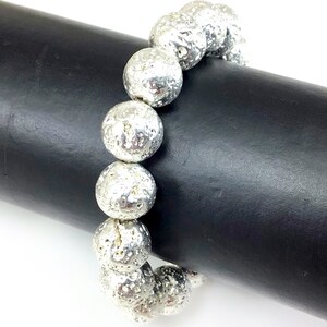 May include: Silver-colored beaded bracelet featuring large, textured, spherical beads. The bracelet is displayed against a black cylindrical background, showcasing its metallic sheen and unique surface details. The beads appear to be approximately 1 inch in diameter.