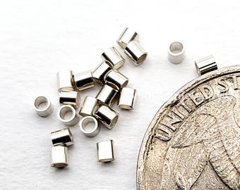May include: A collection of small, shiny, silver-colored metal crimp tubes scattered on a white surface. The tubes are cylindrical with a hole running through the center. A silver coin is partially visible in the image.