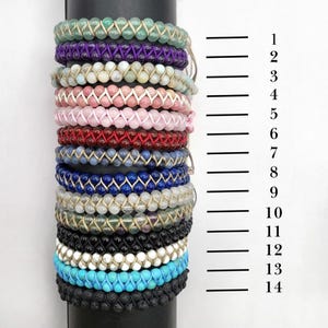 May include: A stack of 14 bracelets made with different colored beads and cords. The bracelets are stacked on a black surface with a ruler beside them. The ruler is marked with numbers 1 through 14.