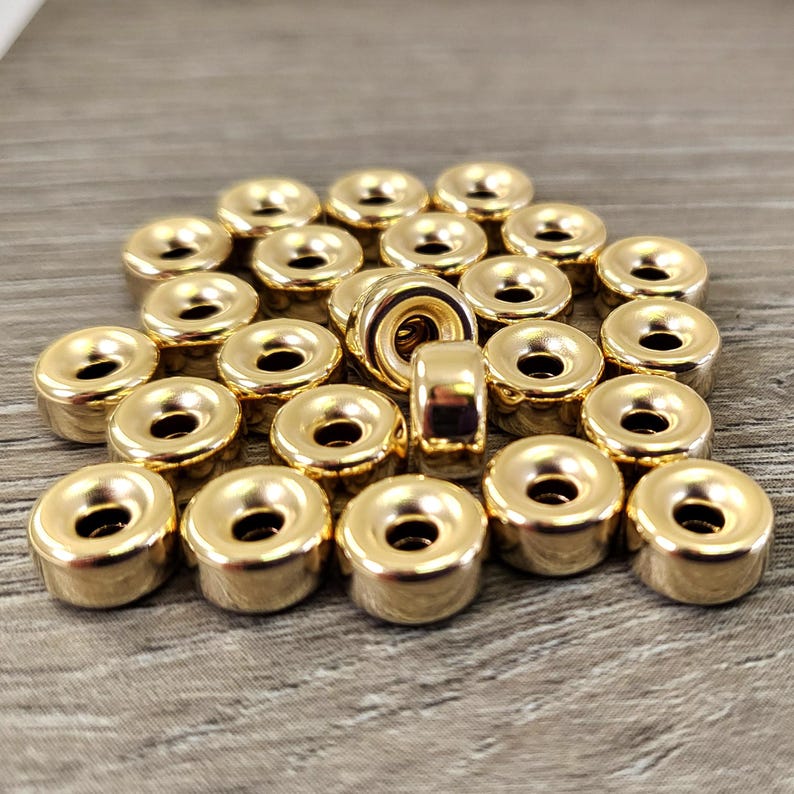Pu&ograve; includere: Un gruppo di 25 perline di metallo dorate con un anello centrale rialzato. Le perline sono disposte in un mucchio su una superficie di legno.
