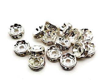 May include: A collection of silver-colored, round beads with a flower-like design. Each bead has a central hole and is adorned with small, sparkling accents. The beads are scattered on a white background, suitable for jewelry making or crafts.