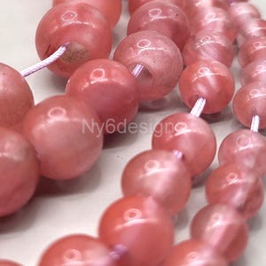 May include: A strand of pink gemstone beads with a light pink, translucent finish. The beads are round and smooth, and they are strung on a white cord.