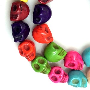 Puede incluir: Una hebra de cuentas de calavera de color turquesa. Las cuentas son de varios colores, incluyendo rojo, naranja, verde, azul y morado.