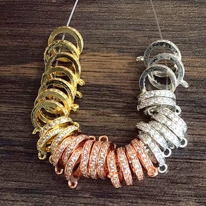 K&ouml;nnte beinhalten: Eine Sammlung von goldenen, silbernen und ros&eacute;goldenen Verschl&uuml;ssen mit klaren Kristall-Akzenten. Die Verschl&uuml;sse sind in einem kreisf&ouml;rmigen Muster gestapelt.
